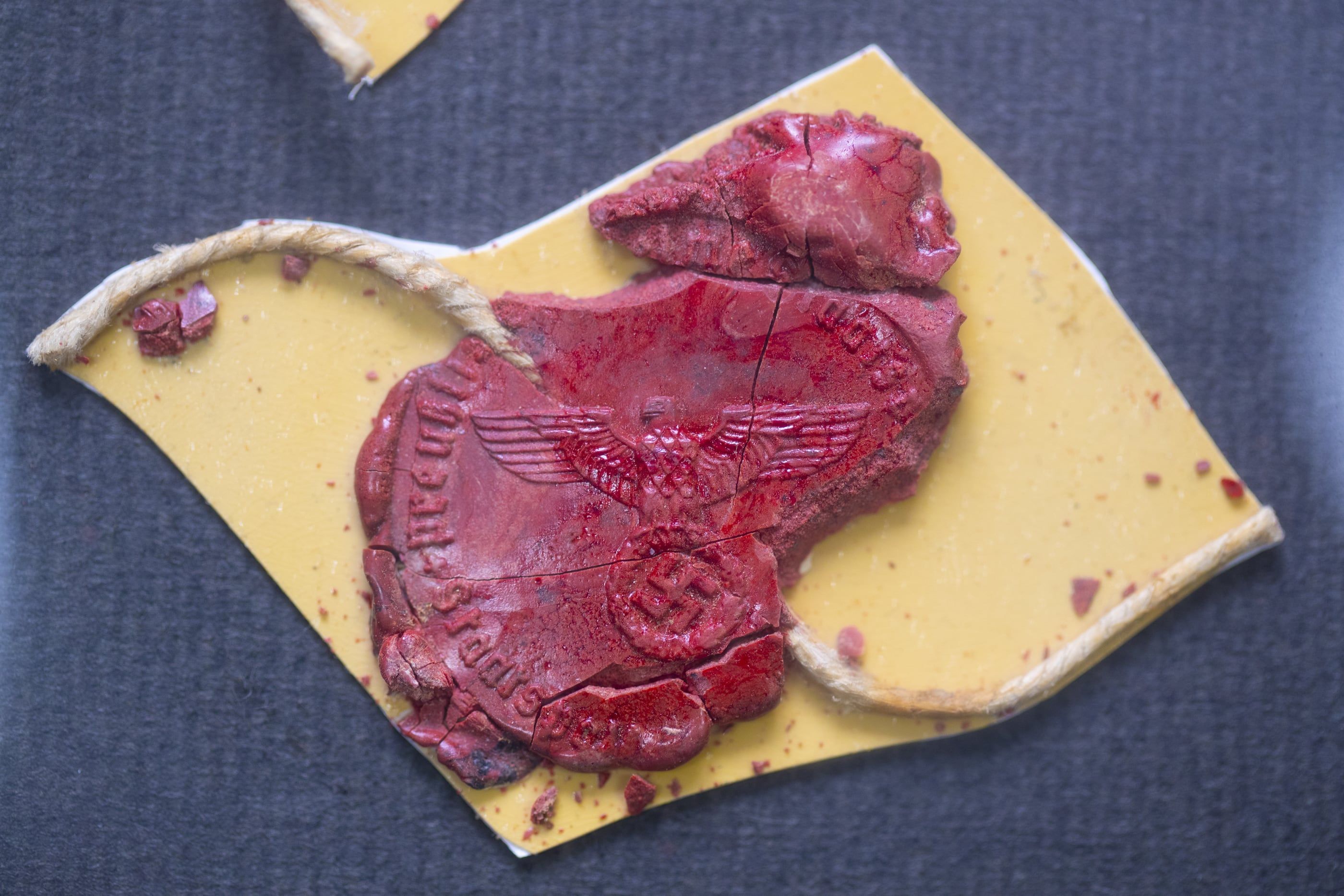 Gebrochenes rotes Lacksiegel mit Reichsadler und Hakenkreuz, mit Siegelschnur, auf einem Stück gelbem Karton