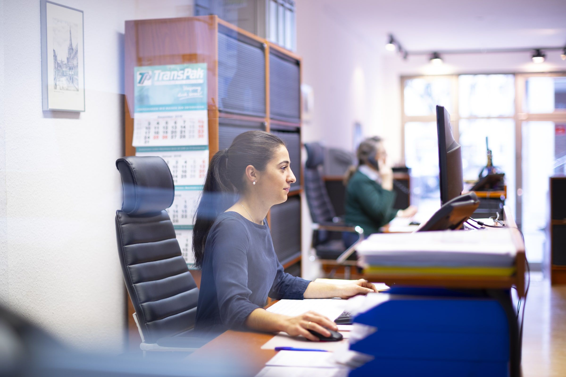 Mitarbeiterin am Schreibtisch im Büro der Druckerei, im Hintergrund eine Kollegin am Telefon