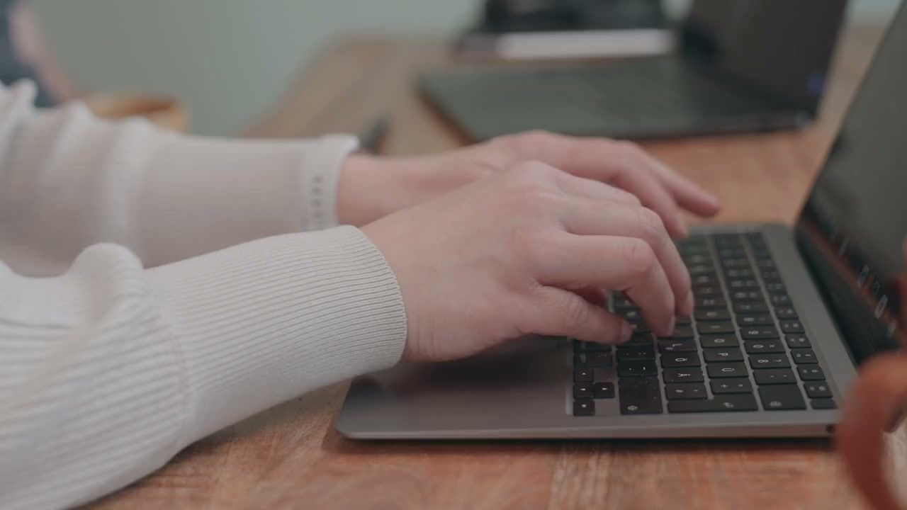 Hände tippen auf der Tastatur eines Laptops, daneben eine orange Tasse